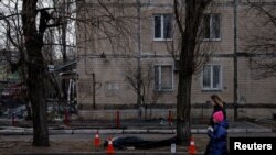 A woman and a child walk past a dead body in front of a residential building that was hit by a shell, as Russia's attack on Ukraine continues, in the Obolon district in Kyiv, Ukraine, March 14, 2022. 
