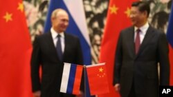 Bendera nasional Rusia dan China berdiri di latar depan saat Presiden Rusia Vladimir Putin, kiri, dan mitranya dari China Xi Jinping berdiri bersama di latar belakang, saat upacara penandatanganan di Wisma Negara Diaoyutai di Beijing, Minggu 9 November 2014. (Foto: AP)