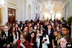 People stand in the Cross Hall of the White House and watch as President Joe Biden speaks during an event to celebrate Equal Pay Day and Women's History Month in the East Room, March 15, 2022.