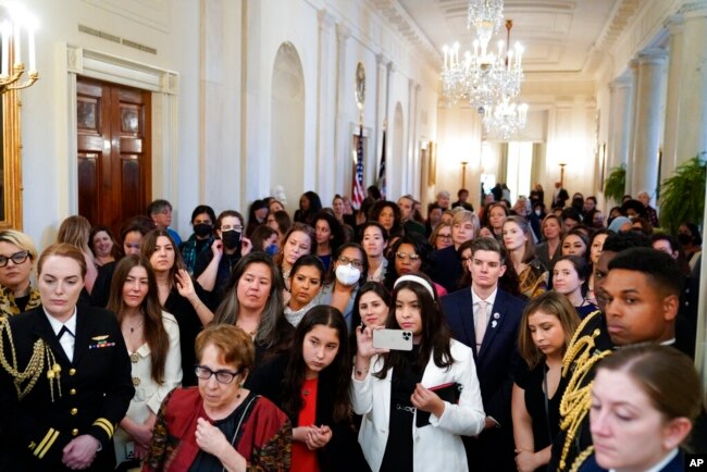 People stand in the Cross Hall of the White House and watch as President Joe Biden speaks during an event to celebrate Equal Pay Day and Women's History Month in the East Room, March 15, 2022.