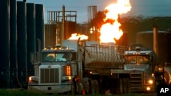 FILE - Drivers and their tanker trucks capable of hauling water and hydraulic fracturing liquid line up near a natural gas burn off flame and storage tanks in Williston, N.D., on June 9, 2014. 