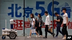 People walk toward a police robot mounted with surveillance cameras patrol past a 5G network advertisement at a shopping district in Beijing, Wednesday, May 15, 2019. U.S. officials listed $300 billion more of Chinese goods for possible tariff hikes…