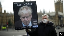 Seorang pengunjuk rasa anti-Partai Konservatif memegang plakat yang menampilkan gambar Perdana Menteri Inggris Boris Johnson dengan tulisan "Now Partygate" yang dilatarbelakangi oleh Gedung Parlemen, di London, Inggris, 8 Desember 2021. (Foto: AP)