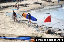 Wisatawan berjalan dengan papan selancar mereka di sepanjang pantai Kuta melewati puing-puing dan sampah yang hanyut oleh air pasang di Bali pada 9 Desember 2018. (Foto: AFP/Sonny Tumbelaka)