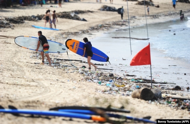 Wisatawan berjalan dengan papan selancar mereka di sepanjang pantai Kuta melewati puing-puing dan sampah yang hanyut oleh air pasang di Bali pada 9 Desember 2018. (Foto: AFP/Sonny Tumbelaka)