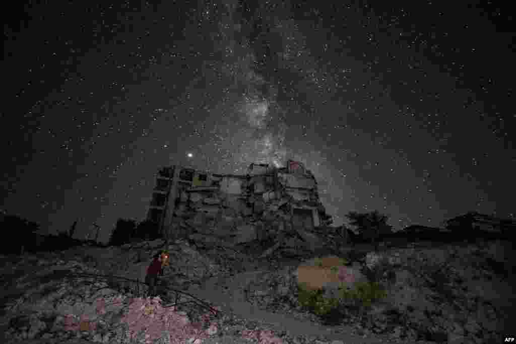 Seorang pria Suriah menikmati malam yang cerah sambil merokok di antara reruntuhan gedung-gedung yang hancur akibat serangan udara di kota Ariha, provinsi Idlib, sementara galaksi Bima Sakti tampak di atas langit.