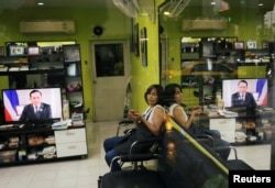 A woman sits on a chair inside a barbershop as Thailand's Prime Minister Prayuth Chan-ocha is seen on a TV screen.