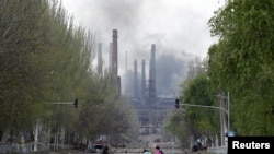 People walk their bikes across the street as smoke rises above a plant of Azovstal Iron and Steel Works during Ukraine-Russia conflict in the southern port city of Mariupol, Ukraine, May 2, 2022.