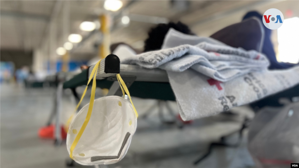 Una mascarilla N-95 cuelga de uno de los catres ubicados en esta bodega convertida en albergue en El Paso para cobijar a los migrantes que son liberados por la Patrulla Fronteriza. Un hombre haitiano descansa en este lugar de paso, donde la mayor parte de las personas se quedan por cortos períodos de tiempo. Cada persona recibe una cobija que ha sido donada por la Cruz Roja. [Foto: Celia Mendoza, mayo 18, 2022, El Paso, Texas]