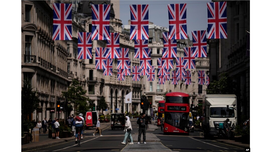 Orang-orang melintasi distrik perbelanjaan Regent Street dengan bendera Union yang tergantung di atasnya untuk menandai Platinum Jubilee yang akan datang dari 70 tahun pemerintahan Ratu Elizabeth II Inggris, di London, 18 Mei 2022. (Foto: AP)