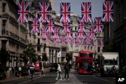 Orang-orang melintasi distrik perbelanjaan Regent Street dengan bendera Union yang tergantung di atasnya untuk menandai Platinum Jubilee yang akan datang dari 70 tahun pemerintahan Ratu Elizabeth II Inggris, di London, 18 Mei 2022. (Foto: AP)