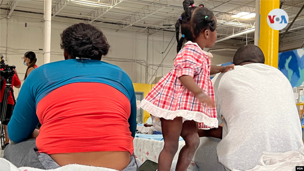 Una niña haitiana se para en uno de los catres de este refugio de paso. Se sostiene de su papá sentado al lado de su pareja. El director de Casa Anunciación, Rubén García, pidió que los rostros de los migrantes no sean mostrados. [Foto: Celia Mendoza. mayo 18, 2022, El Paso, Texas]