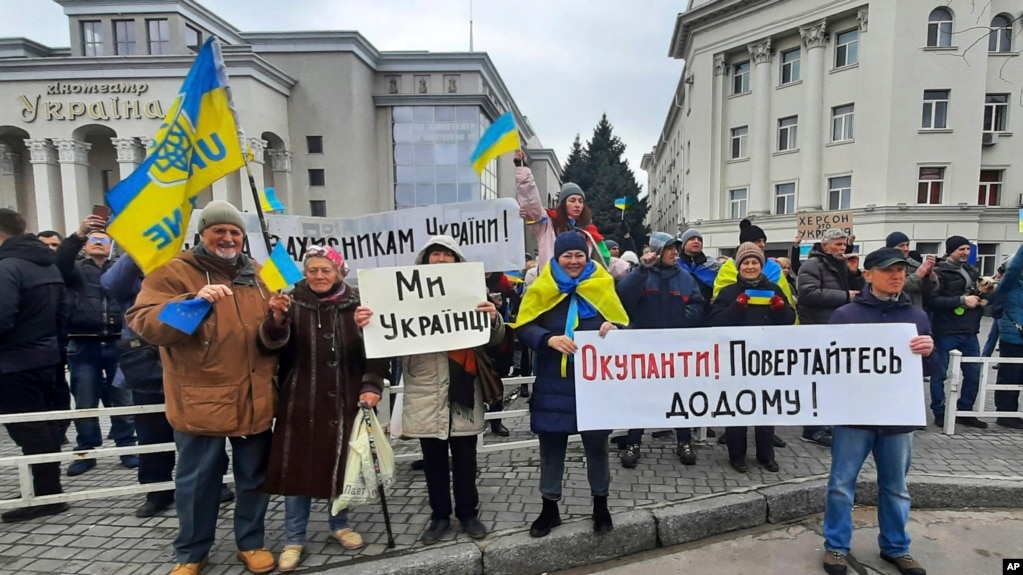 Люди тримають українські прапори та транспаранти з написом «Ми – українці», по центру та «Окупанти! Повертайтеся додому!». під час мітингу проти російської окупації на площі Свободи в Херсоні, Україна, субота, 5 березня 2022 р. (AP Photo/Олександр Чорний)