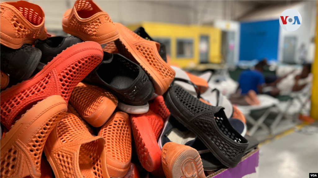 Una caja con zapatos de plástico de varias tallas está a la entrada de este salón en el refugio habilitado por Casa Anunciación, donde los migrantes descansan tras ser puestos en libertad por la Patrulla Fronteriza. [Foto: Celia Mendoza, mayo 18, 2022, El Paso, Texas]