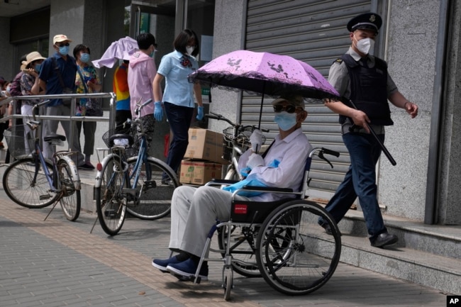 An elderly resident in a wheelchair waits under an umbrella as other elderly residents line up outside a bank to collect their pension as banking services reopen after pandemic measure lockdown are lifted, Tuesday, May 31, 2022, in Beijing. (AP Photo/Ng Han Guan)