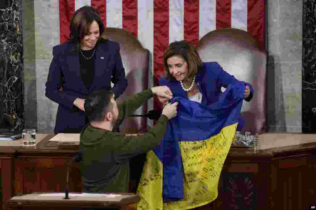 O vice-presidente dos EUA Kamala Harris, à esquerda, e a Presidente da Câmara Nancy Pelosi, à direita, reagem ao Presidente ucraniano Volodymyr Zelenskyy que lhes oferece uma bandeira ucraniana em Washington, 21 de Dezembro de 2022.