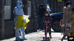 A child wearing a face mask and riding on a scooter passes by a worker in protective suit on his way to collect COVID samples from the lockdown residents in Beijing, Dec. 1, 2022. 