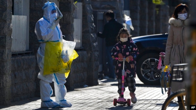 A child wearing a face mask and riding on a scooter passes by a worker in protective suit on his way to collect COVID samples from the lockdown residents in Beijing, Dec. 1, 2022.