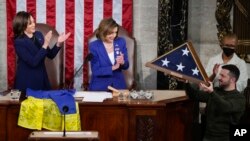 对美国国会参众两院联席会议发表演说的乌克兰总统泽连斯基在副总统哈里斯和众议院议长佩洛西的注视下举着一面赠送给他的美国国旗。他向国会赠送了一面来自前线的乌克兰旗帜。(2022年12月21日)
