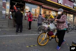 Warga mengenakan masker saat mendorong sepeda dengan barang belanjaannya melewati sebuah toko yang mengontrol arus pembelinya di Beijing, China, Jumat, 25 November 2022. (AP/Ng Han Guan)
