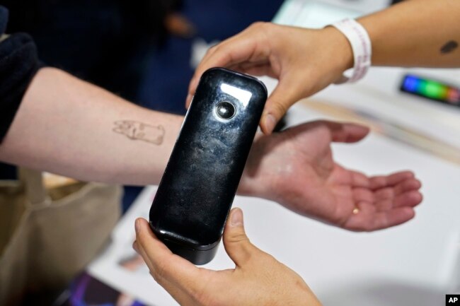 An exhibiter applies a temporary tattoo with a Prinker digital temporary tattoo device during CES Unveiled before the start of the CES tech show, Tuesday, Jan. 3, 2023, in Las Vegas. (AP Photo/John Locher)