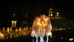 Turis berdiri di depan spanduk raksasa yang menunjukkan topeng emas Raja Tutankhamun di Avenue of Sphinx yang umumnya dikenal sebagai El Kebbash Road di Luxor, Mesir, Jumat, 4 November 2022. (AP/Amr Nabil)