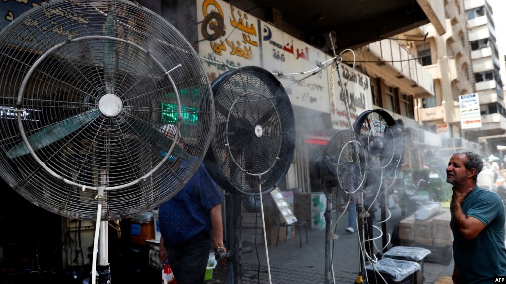 资料照片：2021年6月30日，在伊拉克首都巴格达，一名男子站在风扇旁边，风扇上喷洒着捐助者提供的混合水蒸气，为街道上的行人降温。(photo:VOA)