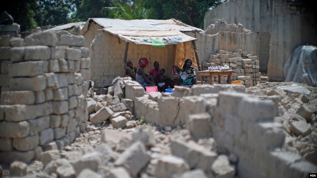A family uses tarps taken from a refugee camp to get shelter from the sun, with bricks for rebuilding their homes all around them in the PK5 neighborhood of Bangui, Feb. 2017. (Z. Baddorf/VOA)