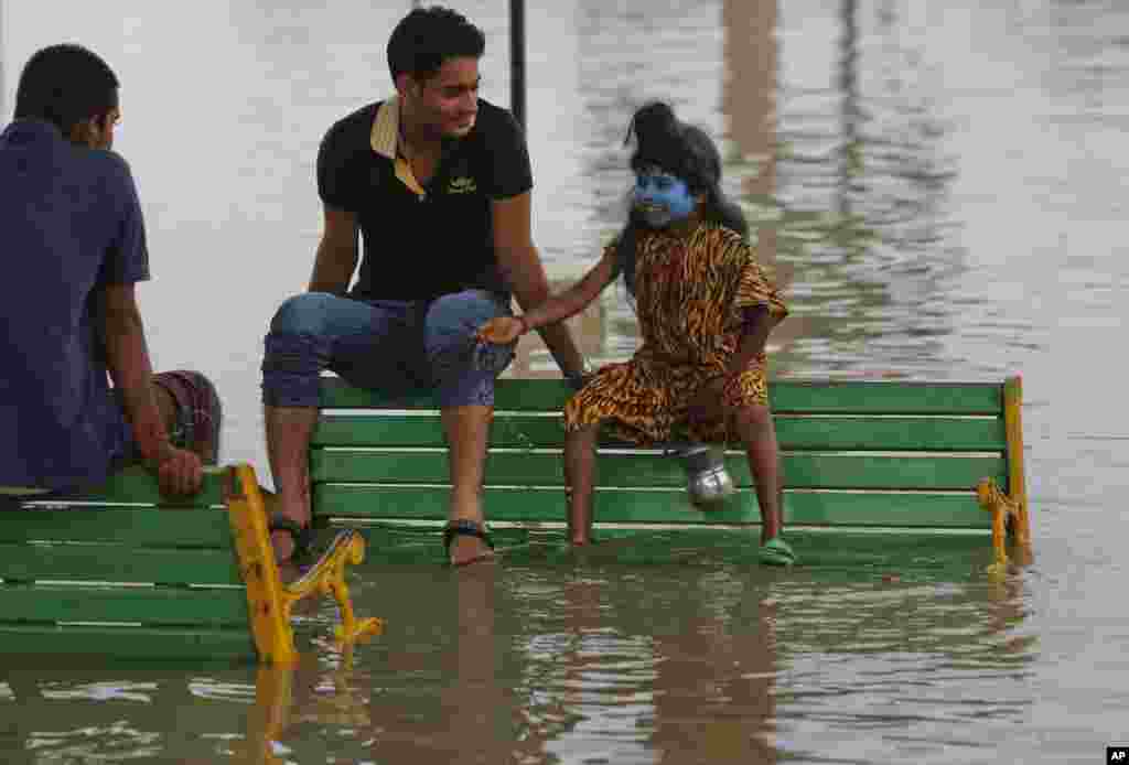 Một cậu bé Ấn Độ ăn mặc như thần Shiva của đạo Hindu trò chuyện với khách du lịch trong khi họ ngồi trên băng ghế bị ngập trong nước lũ trên bờ sông Hằng ở Allahabad.