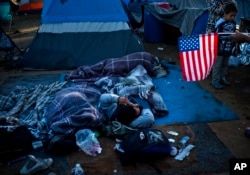 Un migrante que tiene una bandera de los Estados Unidos, a la derecha, habla con otras personas que se despiertan en el Centro Deportivo Benito Juárez que sirve como refugio temporal en Tijuana, México, el lunes 26 de noviembre de 2018.