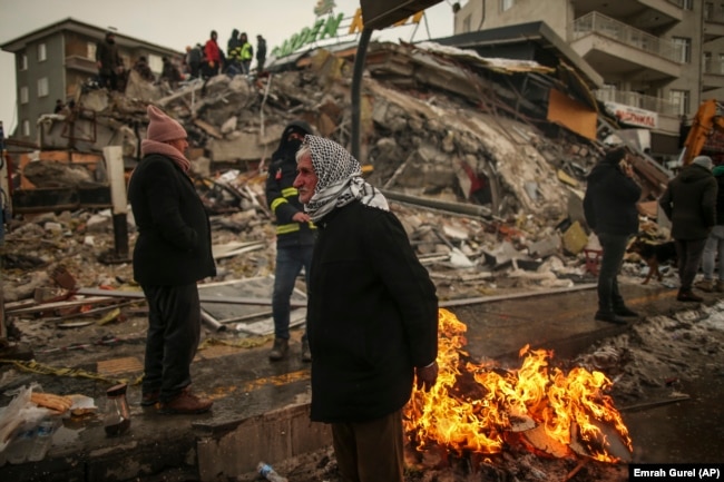 People warm themselves next to a collapsed building in Malatya, Turkey, Tuesday, Feb. 7, 2023. (AP Photo/Emrah Gurel)