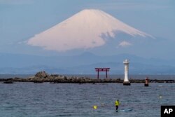 Dengan latar belakang Gunung Fuji, seorang pemain paddleboarder berdiri berlayar di dekat gerbang Torii, gerbang masuk ke kuil Shinto, di Teluk Sagami, 6 April 2022, di Zushi, selatan Tokyo. (Foto: AP)