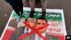 Anti-government protesters stand on a defaced poster with pictures of former Iraqi Prime Minister Nouri al-Maliki, left, and Iraq's Prime Minister-designate Adnan Al-Zurfi, in Tahrir Square, March 18, 2020.