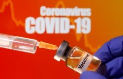 A small bottle labeled with a "Vaccine" sticker is held near a medical syringe in front of displayed "Coronavirus COVID-19" words in this illustration taken April 10, 2020.