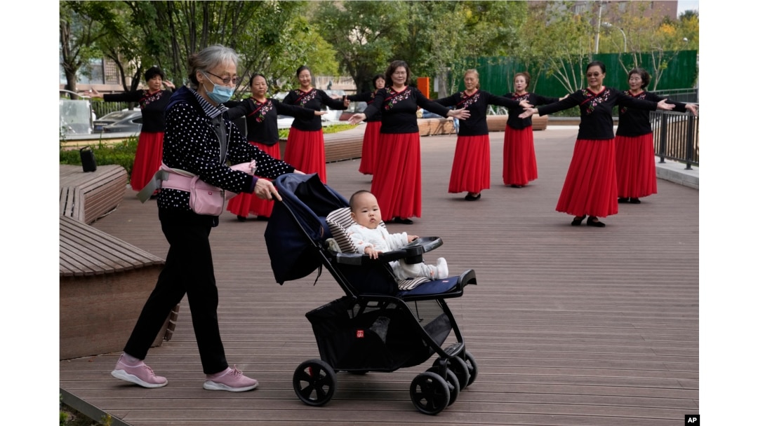 资料照:一名老年妇女推着一个婴儿走过北京一所公园里跳广场舞的妇女。(2021年10月14日)