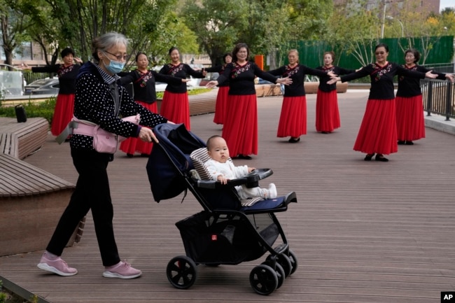 FILE - An elderly woman wearing a mask pushes a baby in a pram past plaza dancers at a park in Beijing, China, Thursday, Oct. 14, 2021. (AP Photo/Ng Han Guan)
