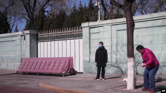 A resident walks her dog near a plainclothes security person standing watch outside the Russian Embassy in Beijing on Tuesday, March 1, 2022. (AP Photo/Ng Han Guan)