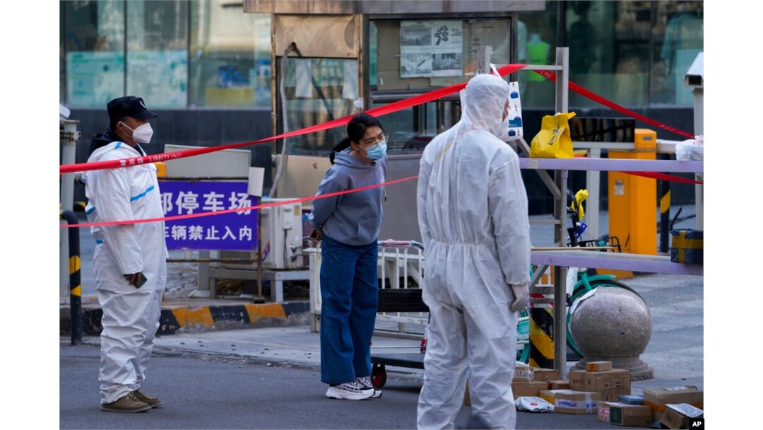 FILE - Security guards wearing protective suits stand watch as a masked woman waits for her parcel at a residential building locked down for health monitoring following a COVID-19 case detected in the area, in Beijing, March 28, 2022.