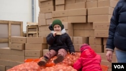 A Ukrainian child eats a slice of bread sitting in a storage facility on sacks of onions as another one plays. Psychiatrists worry about the long-term health effects of the war on Ukraine’s youngsters (Jamie Dettmer/VOA) 