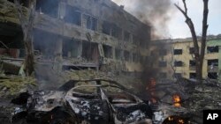 Sebuah mobil terbakar di sisi rumah sakit bersalin yang rusak akibat penembakan di Mariupol, Ukraina, Rabu, 9 Maret 2022. Serangan Rusia telah merusak ebuah rumah sakit bersalin di kota pelabuhan Mariupol yang terkepung, kata pejabat Ukraina. (Foto: AP/Evgeniy Maloletka)