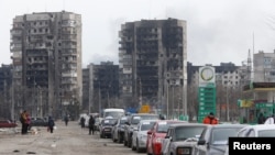 Uma fila de carros com pano de fundo prédios residenciais destruídos pelo conflito Ucrânia-Rússia. Residentes tentam sair da cidade portuária de Mariupol, Ucrânia, 17 Março, 2022.