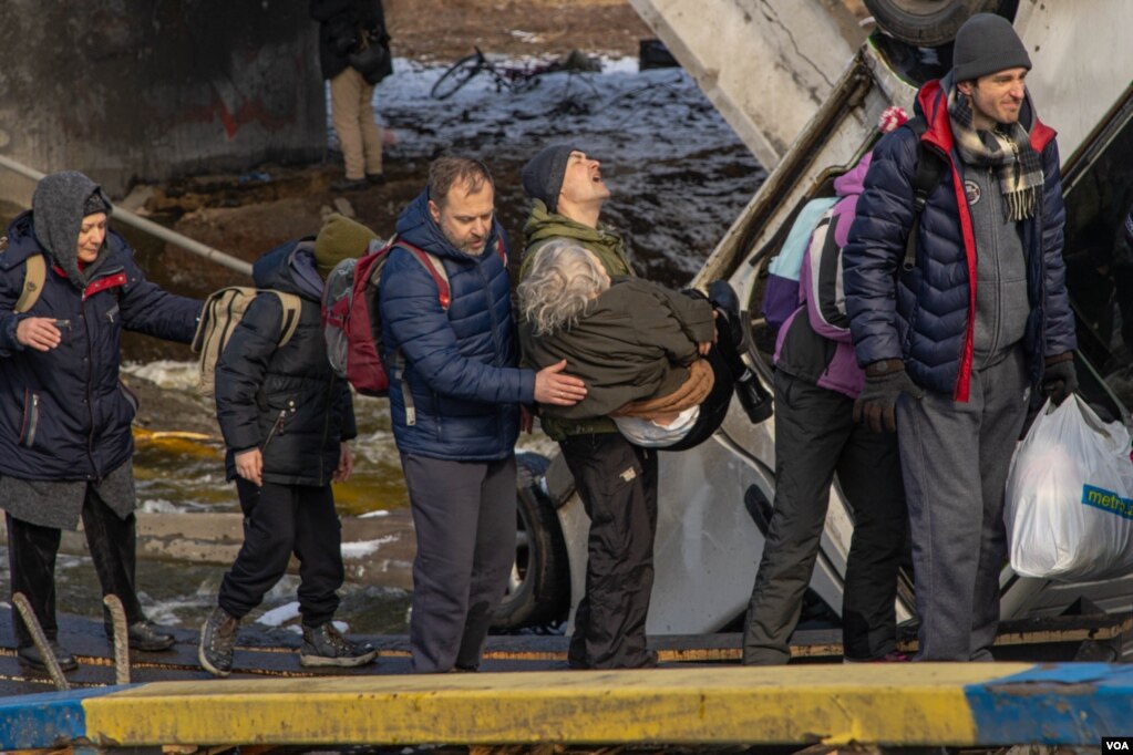 A man carries an elderly woman as a group of people walk through an improvised path created under a bombed bridge over the Irpin River, in Ukraine, March 8, 2022. (Yan Boechat/VOA) 