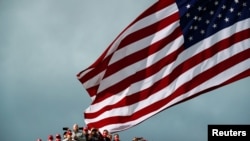 Para pendukung yang mengenakan Topi Make America Great Again terlihat saat bendera AS dikibarkan sebelum dimulainya acara kampanye Presiden AS Donald Trump di Bandara Internasional Des Moines di Des Moines, Iowa, AS, 14 Oktober 2020. (Foto: REUTERS/Carlos Barria )