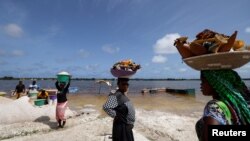 FILE: A worker carries a crate containing newly harvested salt from the edge of Lac Rose also known as Lake Retba as she walks past Female vendors, selling handcrafts for tourists in Niaga, near Dakar, Senegal. Taken Sept. 6, 2022. 