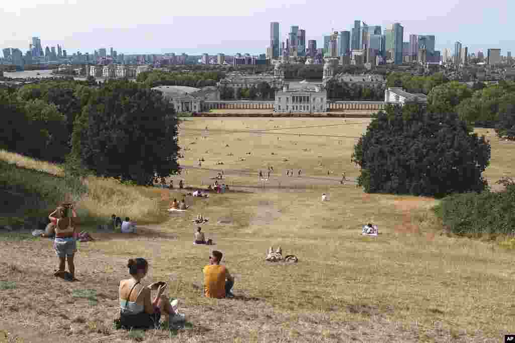 Gente sentada en pasto seco por el sol en el Parque de Greenwich, con el Museo Marítimo y el distrito financiero de Canary Wharf al fondo, en Londres, el 17 de julio de 2022.