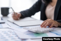 A businesswoman uses a calculator and laptop to work on math finance.