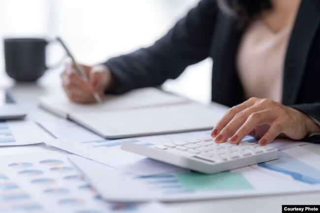 A businesswoman uses a calculator and laptop to work on math finance.