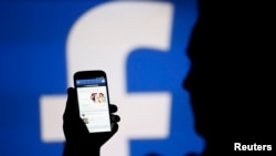 FILE - A man is silhouetted against a video screen with an Facebook logo as he poses with a smartphone.