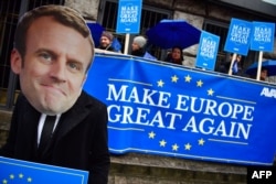Activists of the Avaaz civic organization wear a mask of French President Emmanuel Macron and hold up posters and a banner reading "Make Europe Great Again."