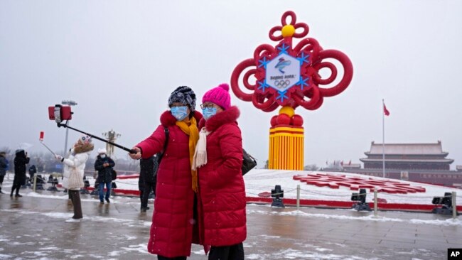 Gratë që mbajnë maska për fytyrën të printuara me një slogan për Lojërat Olimpike Dimërore të Pekinit bëjnë një selfie me një dekorim të lojërave në sfond, në sheshin Tiananmen në Pekin, 20 janar 2022 (AP Photo/Andy Wong)